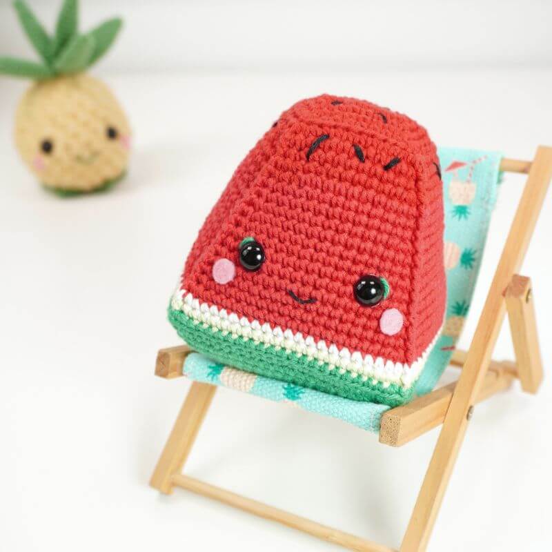 Amigurumi de sandía hecho a mano con hilo rojo, verde y blanco y patrón de piña de crochet al fondo